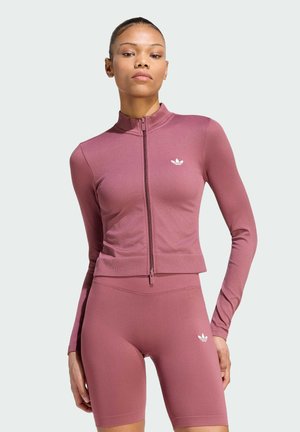 Mujer con chaqueta ajustada de cremallera color malva y shorts ciclistas a juego de cintura alta con logos blancos de Adidas en el pecho y el muslo.
