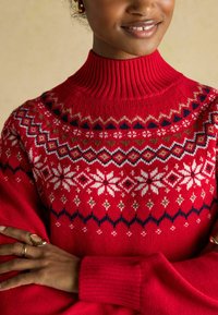 Red knitted sweater with a high ribbed collar, featuring a white snowflake and geometric pattern on the yoke. Soft texture, short sleeves.