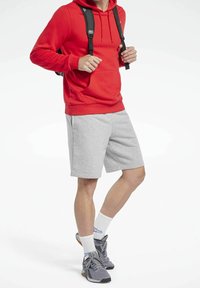 Sweat-shirt à capuche rouge, shorts gris, chaussures de sport grises, chaussettes blanches. Les bretelles du sac à dos sont visibles. Tenue décontractée avec un mélange de couleurs et de textures.