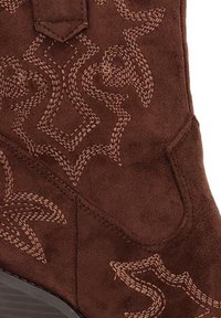 Bota de ante marrón con intrincadas costuras en tonos beige formando patrones geométricos y florales. Textura suave con punta afilada y suela resistente.