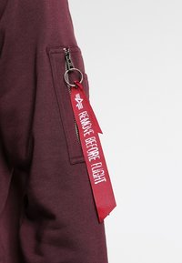 Bordeauxrotes Sweatshirt mit einer seitlichen Reißverschlusstasche und einem roten Stoffetikett mit der Aufschrift "REMOVE BEFORE FLIGHT", versehen mit einem metallischen Ringakzent.