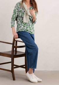 Grünes Oberteil mit floralem Muster aus leichtem Stoff, kombiniert mit lässigen Denim-Jeans und weißen Turnschuhen, sitzt auf einem Holzstuhl.