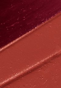 Prodotti per labbra opachi in tonalità rosso scuro e terracotta, con una texture liscia e cremosa e una finitura leggermente lucida, che mostrano un'applicazione strati.