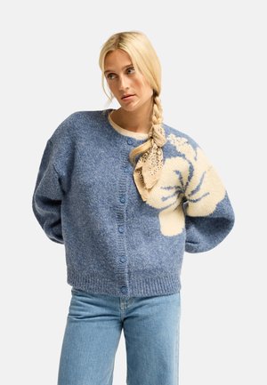 Femme avec une tresse blonde attachée avec un foulard beige, portant un cardigan bleu boutonné à grand motif floral crème et un jean bleu clair.