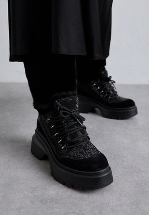 Personne portant des bottes à plateforme noires en tissu texturé avec des lacets, debout sur un sol gris, vêtue de noir.