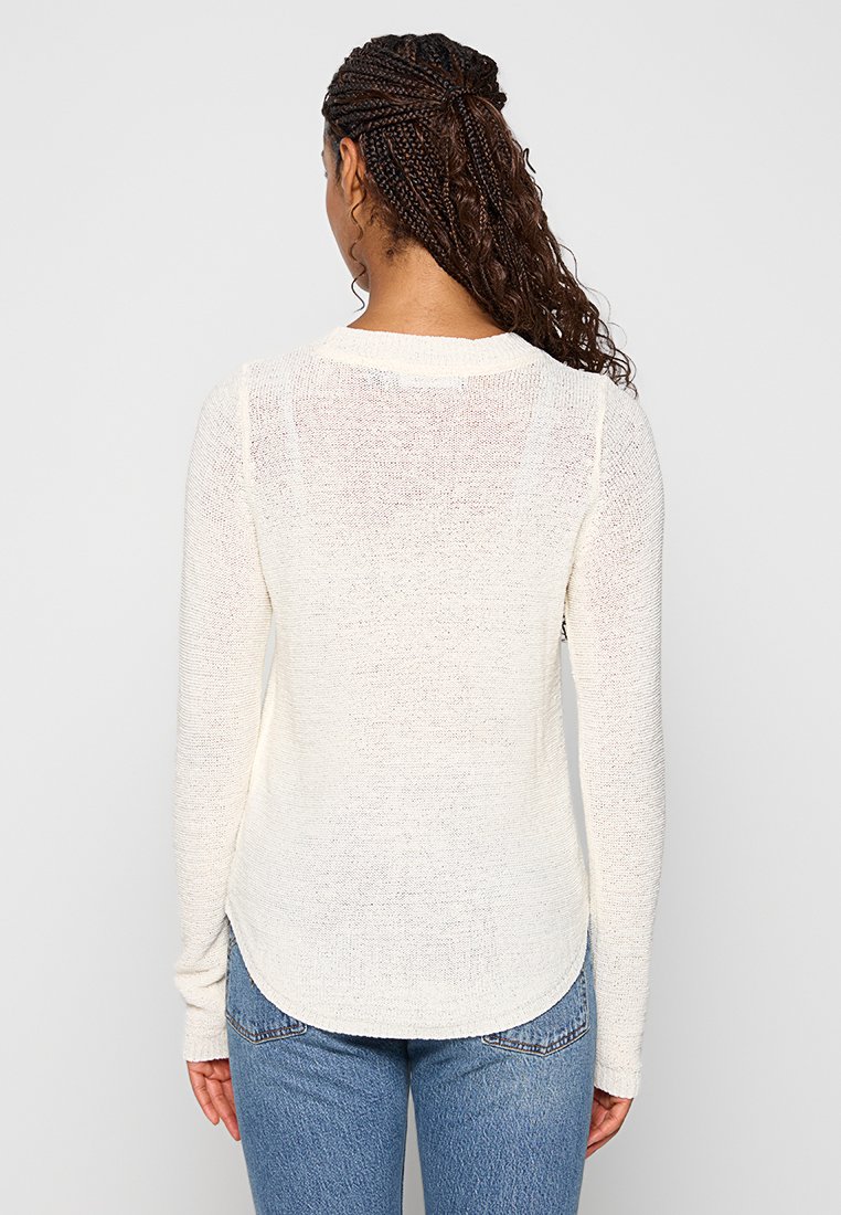 Biały sweter z długim rękawem, o zaokrąglonym dole, miękkiej teksturze i lekko prześwitującym wzorze. W komplecie z niebieskimi dżinsami.