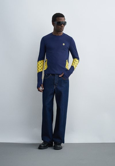 Άνδρας φοράει μεγάλα σκουρόχρωμα γυαλιά ηλίου, μπλε σκούρο πουκάμισο με κίτρινα σχέδια σε σχήμα ημισελήνου στα μανίκια, σκούρα τζιν παντελόνια με φαρδιές φάσες και μαύρα παπούτσια.