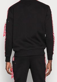 Felpa nera con strisce laterali rosse e testo, polsini a coste e tasca con zip sulla manica sinistra. Tessuto morbido.