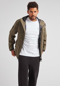 Giacca impermeabile verde oliva con cappuccio nero, multiple tasche frontali e chiusura con zip, indossata sopra una maglietta bianca e pantaloni scuri.
