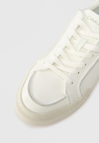 Adidași din piele albă cu vârf rotunjit, design cu șireturi, accent gri subtil pe margine și talpă din cauciuc texturat. Numele brandului este vizibil în interior.