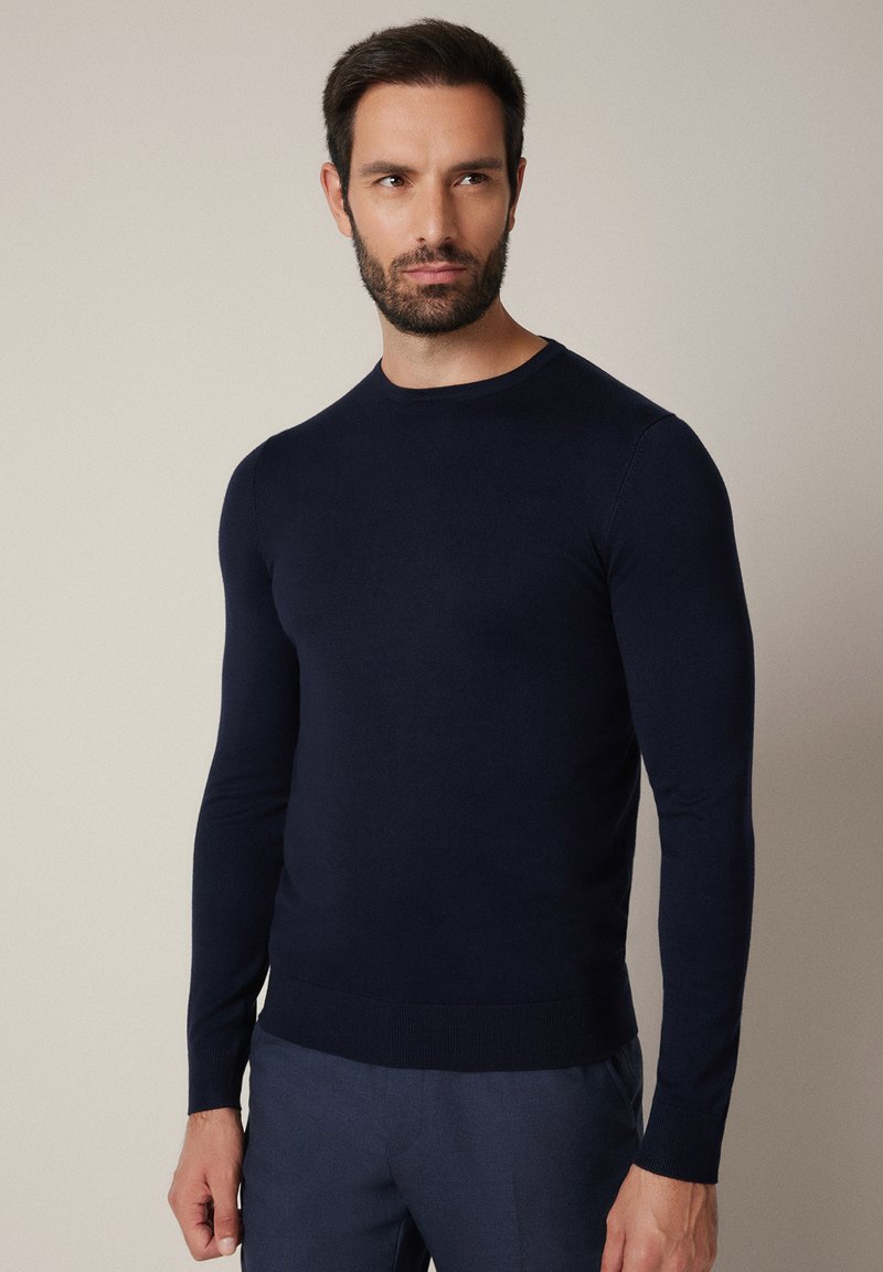 Granatowy sweter z długim rękawem i okrągłym dekoltem, wykonany z gładkiego materiału. Posiada ściągacze na mankietach i dole oraz dopasowany krój.