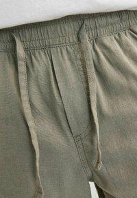 Olivgrüne Jogginghosen mit elastischem Bund, Zugband und sichtbaren Nähten. Der Stoff hat eine weiche Textur und einen leichten Glanz.