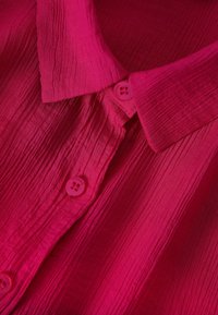 Camicia fucsia con colletto e una texture arricciata, caratterizzata da bottoni rosa lungo la patta, che mette in risalto un tessuto liscio e leggero.