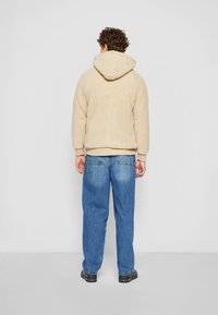 Beige fleece kapucnis pulóver, texturált felülettel, kapucnival és bordázott aljjal, világoskék laza farmerral és fekete cipővel párosítva.