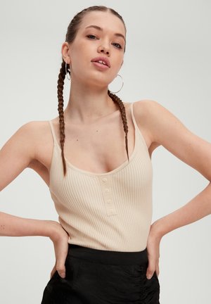 Beige geribbelde tanktop met dunne spaghetti bandjes, met een ronde halslijn en drie knopen aan de voorkant; gedragen met een zwarte rok.