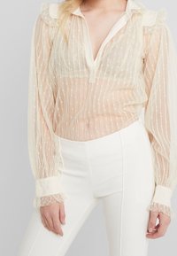 Przezroczysta, kość słoniowa bluzka z pionowymi paskami, falbanianym kołnierzem i mankietami oraz delikatnym kropkowanym wzorem; zestawiona z dopasowanymi białymi spodniami.