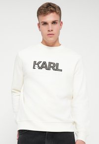 Mladý muž v bielom mikinovom saku Karl Lagerfeld s čiernym textom, stojaci na čistom bielom pozadí.