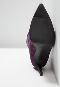 Chaussure à talon haut en daim violet avec un bout pointu et une semelle noire texturée, reposant sur un fond clair.