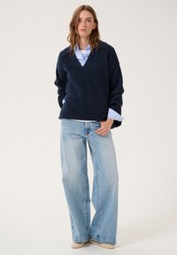 Pulover bleumarin cu decolteu în V, cu mâneci lungi, purtat peste o cămașă albastră deschisă și blugi cu croi larg din denim deschis. Textură moale din tricot și croi lejer.
