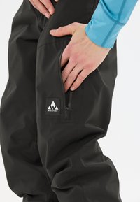 Schwarze wasserdichte Hosen mit einer Reißverschlusstasche am Oberschenkel, mit dem "Whistler"-Logo in Weiß auf schwarzem Hintergrund, glatte Textur.