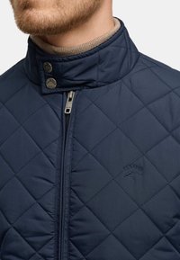 Navy gesteppte Jacke mit einem hohen Kragen, Druckknöpfen und einem Reißverschluss vorne. Weicher Innenstoff mit einem gestickten Logo-Detail.