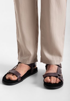 Rudos odinės šlepetės su reguliuojamais dirželiais, atvira pirštų dizaino, tekstūruota pėdų dalis ir juodas guminis padas, derinamos su šviesiai bežė kelnėmis.