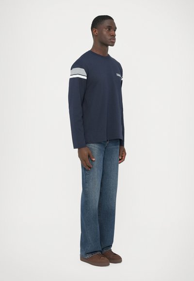 Μπλε σκούρο μπλουζάκι με μακριά μανίκια και γκρι-λευκές ρίγες στα μανίκια, σε συνδυασμό με ανοιχτό μπλε τζιν με ίσια γραμμή και καφέ παπούτσια.