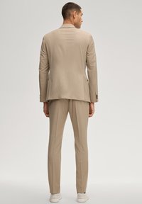 Mann steht mit dem Rücken zur Kamera, trägt einen beigen Anzug mit passenden Hosen und weißen Sneakers, einfarbiger hellgrauer Hintergrund.