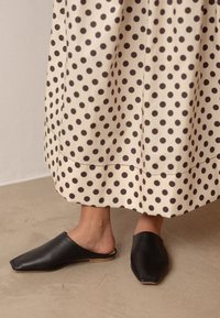 Juodos odinės mūlos su smailiu pirštu ir atvira nugara, derinamos su grietinėlės spalvos sijonu, puoštus dideliais rudais taškais. Lygus paviršius ir minimalistinis dizainas.