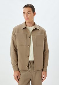 Beige katoenen jack met een kraag en twee grote borstzakken. Knopen aan de voorkant en lange mouwen. Gecombineerd met bijpassende lichtgekleurde broek.