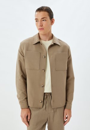 Chaqueta de algodón color beige con cuello y dos grandes bolsillos en el pecho. Cierre frontal de botones y mangas largas. Combinada con pantalones de un color claro a juego.