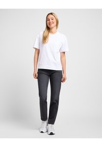 Camiseta blanca de algodón con un bolsillo en el pecho, combinada con jeans negros. El modelo lleva zapatillas deportivas blancas con cordones y una suela texturizada.