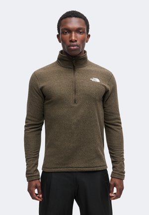 Voksen mand iført en olivengrøn fleece med kvart-lynlås og The North Face-logo på brystet, stående foran en ensfarvet lys baggrund.