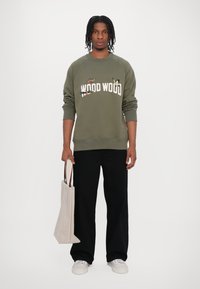 Olivovo zelená mikina so textom "WOOD WOOD" a vyšívanou motýľom, v kombinácii s čiernymi širokými nohavicami a béžovou taškou. Biele tenisky.