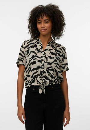 Frau mit lockigem Haar, die ein kurzärmeliges schwarzes und beiges Tiermusterhemd trägt, das in der Taille gebunden ist, und hochtaillierte schwarze Hosen.