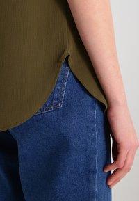 Bras et main à côté de la poche d'un jean bleu, chemise olive verte texturée avec une fente sur le côté.