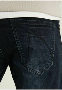 Jeans di denim scuro con un design distintivo della tasca posteriore, caratterizzato da un accento triangolare. Il tessuto appare leggermente texturizzato e consumato.