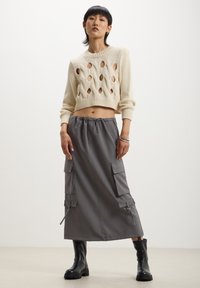 Maglione a maglia crema con ritagli a diamante abbinato a una gonna cargo grigia, lunga fino al ginocchio, con tasche laterali e vita elastica. Stivali neri.