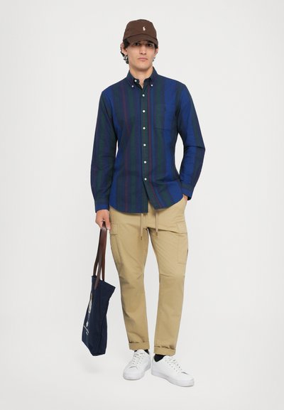 Camisa de botones a rayas en azul oscuro y verde, pantalones cargo beige, gorra marrón, zapatillas blancas y una bolsa tote azul marino con un detalle de diseño.