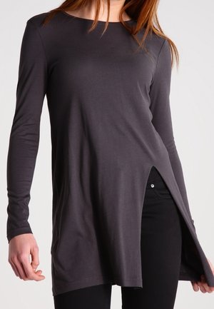 Frau trägt ein langärmeliges, dunkelgraues Tunika-Oberteil mit einem Schlitz an der Vorderseite, kombiniert mit schwarzer Hose vor einfachem Hintergrund.