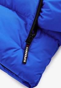 Sinine puffer-jope, millel on sile ja läikiv tekstuur, must tõmblukk ja kaubamärgiga silt "SUPERDRY".