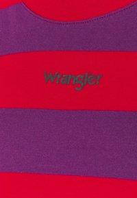 Röd och lila randig tyg med svart Wrangler-logotyp centrerad på bröstområdet av en skjorta.