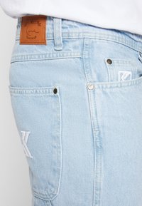 Ljust blå denimjeans med vit broderad "K"-logotyp på bakfickan och läderpatch i brunt med varumärkesnamnet på midjan.