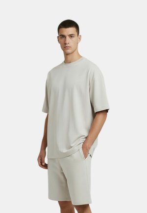 Giovane uomo che indossa una camicia leggera grigio chiaro a maniche corte e pantaloncini abbinati fino al ginocchio, in piedi con una mano in tasca.