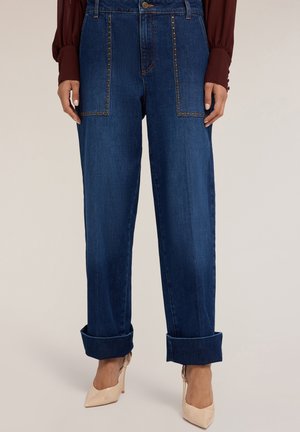 Vaqueros de mezclilla en azul oscuro con un corte holgado, con bolsillos remachados y puños enrollados. Se complementan con zapatos de tacón alto nude.