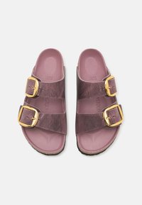 To brune-rosa sandaler med to brede remmer, hver med gullspenner. Sålen er teksturert og mørk med en formet fotseng.