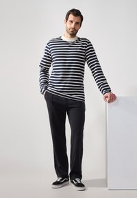 Ein Mann steht neben einem weißen Podest, trägt ein navy- und weiß gestreiftes Langarmshirt, schwarze Hose und schwarze Sneakers, vor einem neutralen Hintergrund.