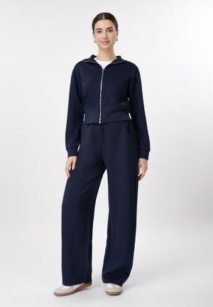 Nő áll, tengerészkék cipzáras dzsekit, hozzá illő bő szárú nadrágot, fehér inget és fehér sportcipőt visel, fehér háttér előtt.