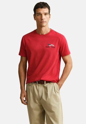 Mann trägt ein rotes T-Shirt mit einer kleinen US-Flagge und dem Schriftzug "GANT 1949", kombiniert mit beigen Hosen und einem braunen Gürtel, steht mit einer Hand in der Tasche.