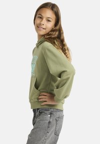 Jeune fille aux cheveux longs et ondulés portant un sweat à capuche vert et un jean gris, debout avec une main sur la hanche, souriant légèrement.